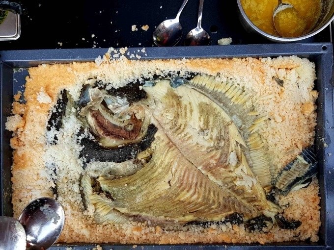 Baked fish in a salt crust on a tray, surrounded by spoons and a bowl of sauce. The fish is partially exposed., &copy; Kultweine-Kochwelten-Kunst&uuml;cke - Kochschule Sonja Lenz