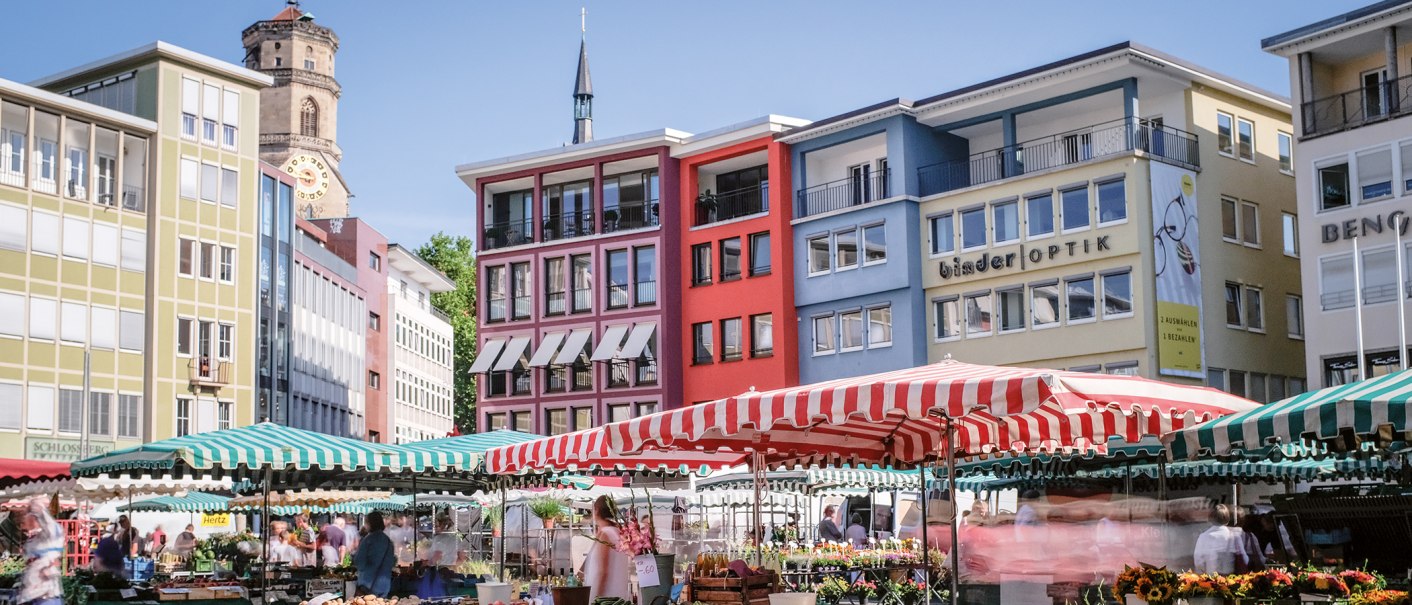 Wochenmarkt in Stuttgart mit bunten Marktständen und Gebäuden im Hintergrund. Menschen schlendern zwischen den Ständen, während die Sonne scheint., © SMG, trickytine Wochenmarkt in Stuttgart mit bunten Marktständen und Gebäuden im Hintergrund. Menschen schlendern zwischen den Ständen, während die Sonne scheint., © SMG, trickytine