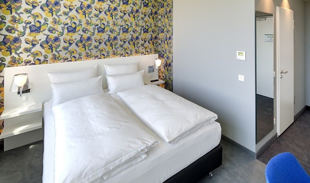 Modern hotel room with double bed, colorful wallpaper and gray walls. A blue chair stands in the foreground., © Hotel am Remspark Modern hotel room with double bed, colorful wallpaper and gray walls. A blue chair stands in the foreground., © Hotel am Remspark