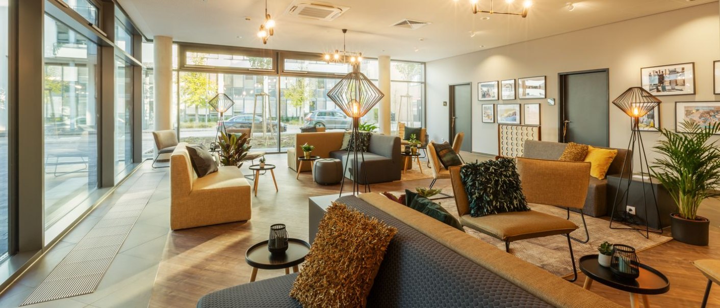 Moderne Lobby mit gemütlichen Sitzgelegenheiten, Pflanzen und großen Fenstern. Dezente Beleuchtung und Wanddekorationen schaffen eine einladende Atmosphäre., © Novum Hospitality