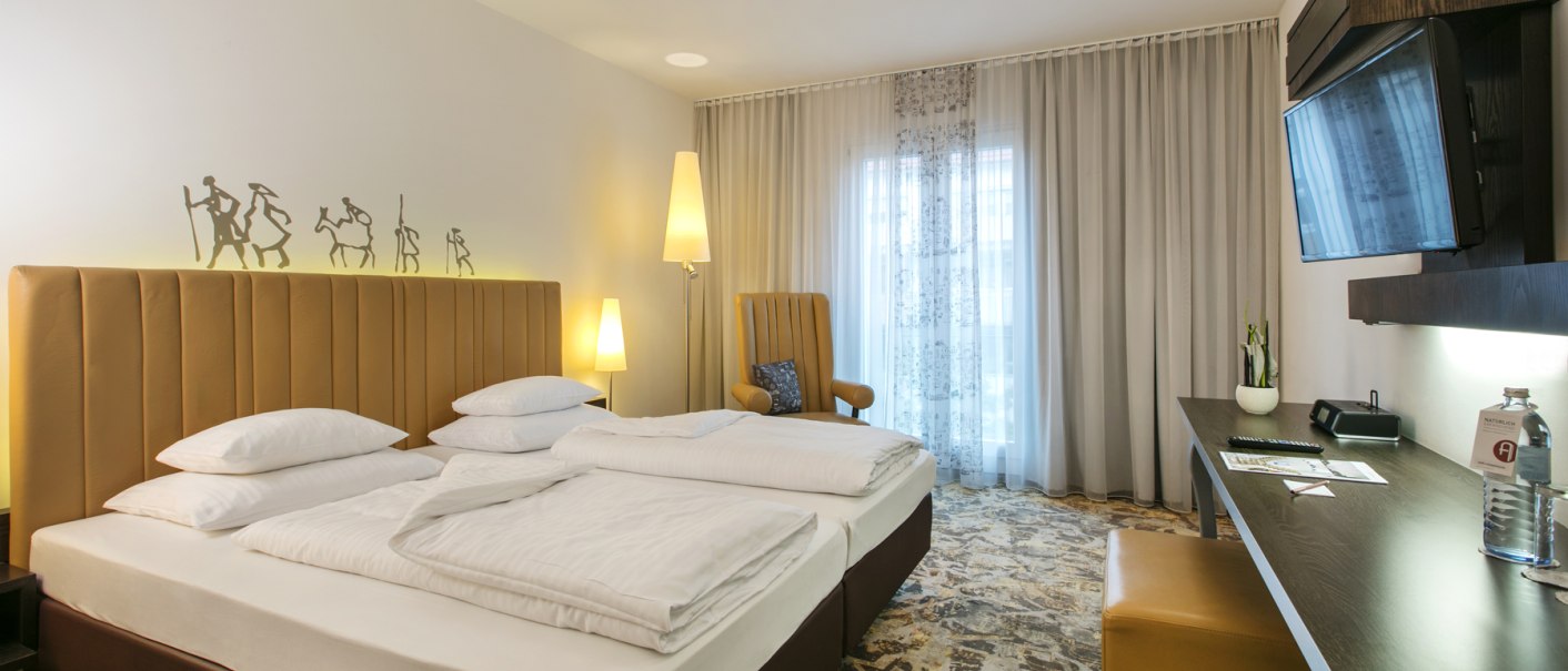 Modern hotel room with double bed, desk, TV and armchair. Subtle wall decoration and large windows with curtains., © ARCOTEL Hotels Modern hotel room with double bed, desk, TV and armchair. Subtle wall decoration and large windows with curtains., © ARCOTEL Hotels
