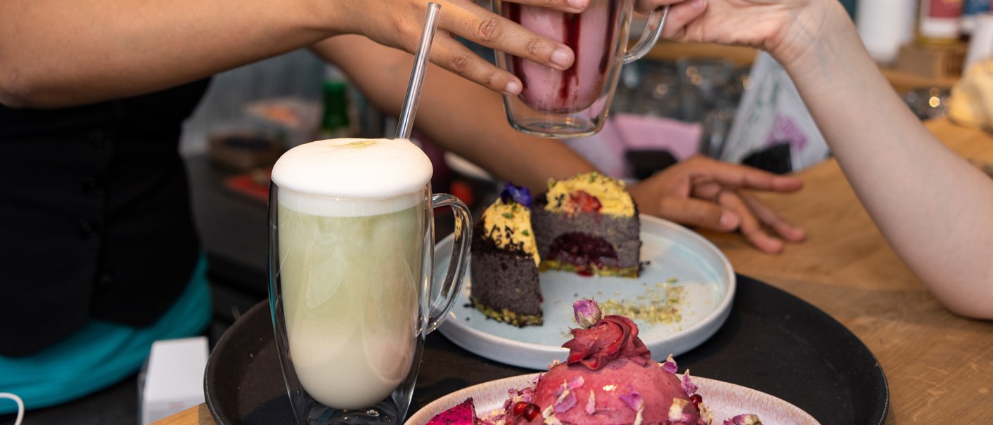 Zwei Personen stoßen mit Getränken an, während auf dem Tisch Kuchen serviert wird. Ein Getränk hat einen Schaum, der Kuchen ist dekorativ verziert., © SMG Zwei Personen stoßen mit Getränken an, während auf dem Tisch Kuchen serviert wird. Ein Getränk hat einen Schaum, der Kuchen ist dekorativ verziert., © SMG