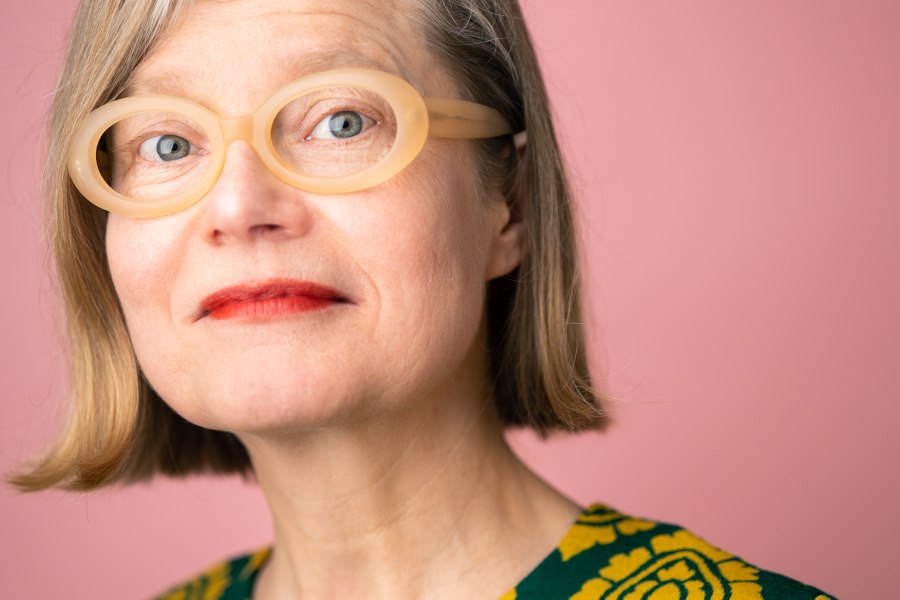 Eine Frau mit blonden Haaren und auffälliger, runder Brille blickt in die Kamera. Der Hintergrund ist rosa, ihr Oberteil hat ein gelbes Muster., © Foto: Annette Koroll Eine Frau mit blonden Haaren und auffälliger, runder Brille blickt in die Kamera. Der Hintergrund ist rosa, ihr Oberteil hat ein gelbes Muster., © Foto: Annette Koroll