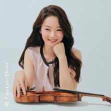 Eine Frau mit langen Haaren lächelt in die Kamera und stützt ihren Kopf auf die Hand. Vor ihr liegt eine Geige auf dem Tisch., © links im Bild Eine Frau mit langen Haaren lächelt in die Kamera und stützt ihren Kopf auf die Hand. Vor ihr liegt eine Geige auf dem Tisch., © links im Bild