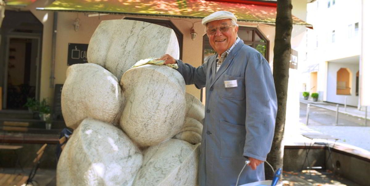 Ein älterer Mann in blauer Arbeitskleidung reinigt eine Steinskulptur. Er trägt eine Mütze und hält einen Eimer in der Hand. Im Hintergrund ist ein Gebäude., © Stuttgart-Marketing GmbH Ein älterer Mann in blauer Arbeitskleidung reinigt eine Steinskulptur. Er trägt eine Mütze und hält einen Eimer in der Hand. Im Hintergrund ist ein Gebäude., © Stuttgart-Marketing GmbH