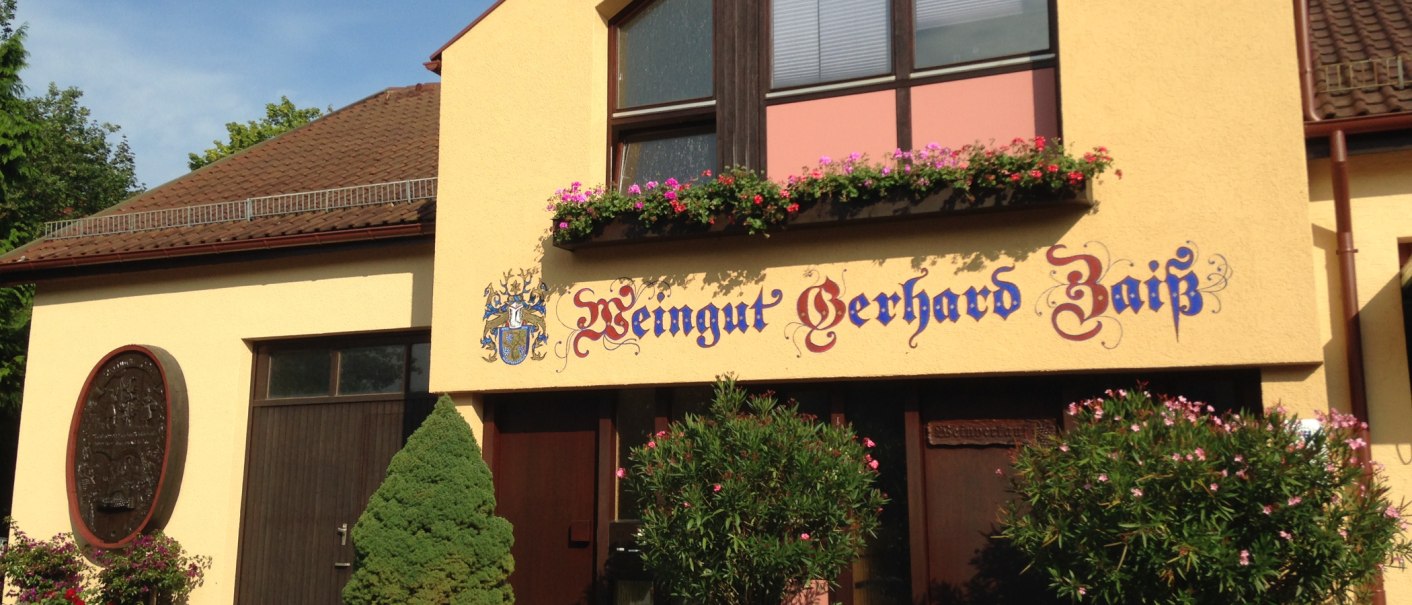 Gelbes Gebäude mit der Aufschrift 'Weingut Gerhard Zaiß', umgeben von Blumen und Pflanzen., © Simone Mack