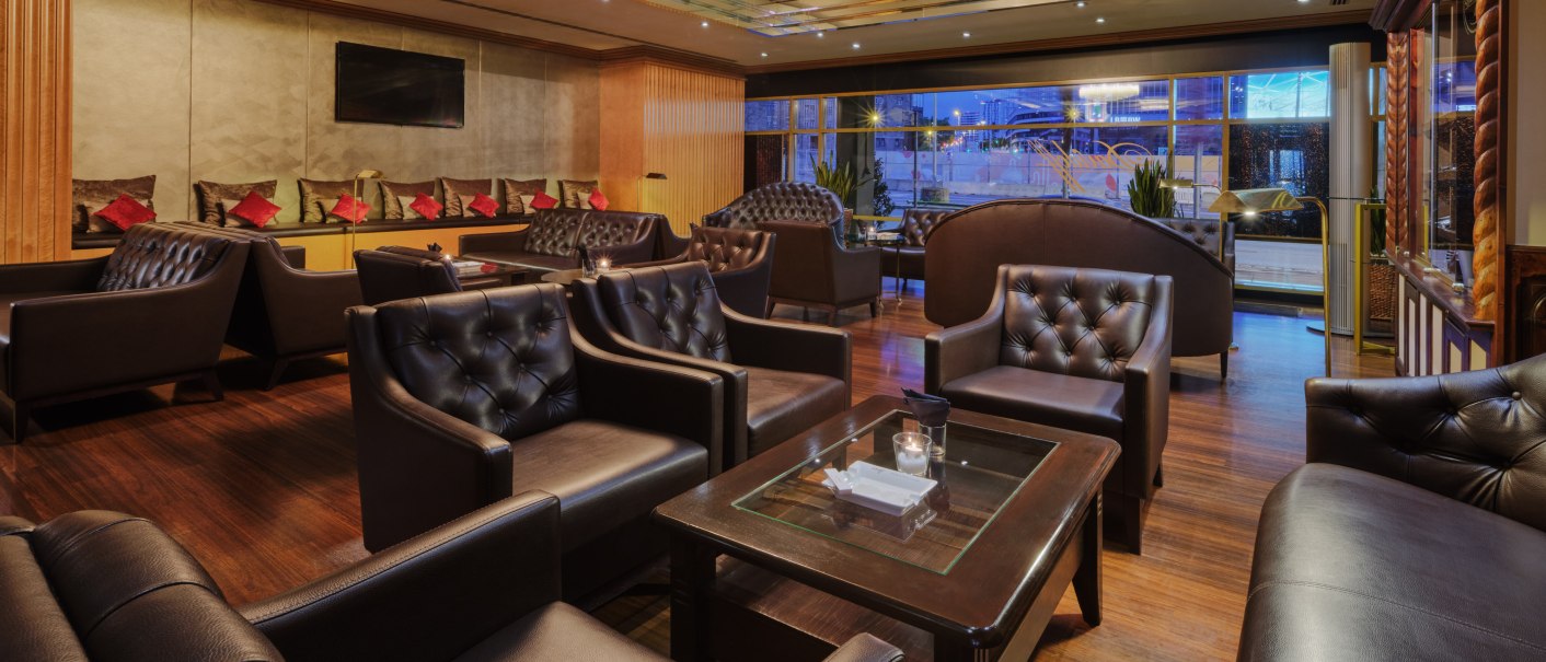 Elegant lounge with dark leather armchairs, wooden floor and a chandelier. Large windows offer a view, red cushions set the tone., &copy; Steigenberger Graf Zeppelin