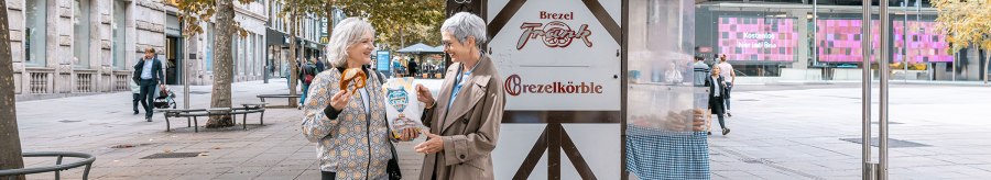 Two women are laughing in front of a pretzel stand on Schlossplatz. One is holding a pretzel, the other a bag. Autumnal trees in the background., © SMG, Sarah Schmid Two women are laughing in front of a pretzel stand on Schlossplatz. One is holding a pretzel, the other a bag. Autumnal trees in the background., © SMG, Sarah Schmid