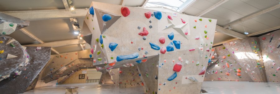 Innenansicht der VELS Boulderhalle mit Kletterwänden und bunten Griffen unter einem gewölbten Dach., © Boulderhalle Vels