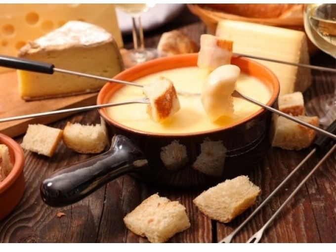 Ein Fondue-Topf mit geschmolzenem K&auml;se, umgeben von Brotst&uuml;cken und K&auml;sebl&ouml;cken auf einem Holztisch., &copy; Cool-Tours StattReisen