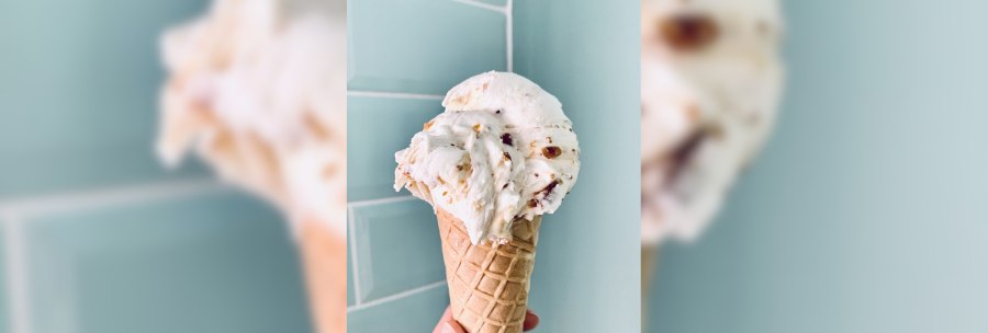 A hand holds an ice cream cone in front of a blue tiled wall. The ice cream has a creamy texture with pieces of nuts., © Zur Schleckerei A hand holds an ice cream cone in front of a blue tiled wall. The ice cream has a creamy texture with pieces of nuts., © Zur Schleckerei