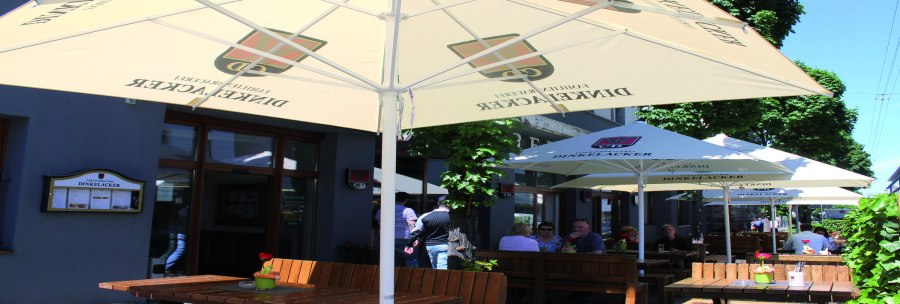 Outdoor area of the Dinkelacker brewery restaurant with wooden benches and parasols. Guests sit in the shade and enjoy the weather., © © Stuttgart-Marketing GmbH (SMG)