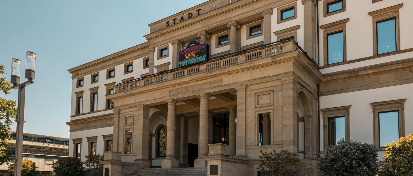 Das StadtPalais mit klassischer Fassade, Säulen und breiter Treppe, unter blauem Himmel. Ein Schild mit bunter Aufschrift ist sichtbar., © Stuttgart Marketing GmbH, Sarah Schmid