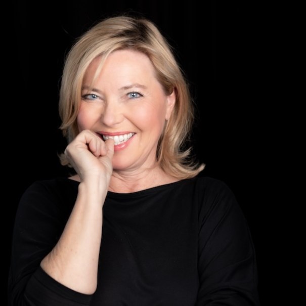 A woman with blonde hair smiles in a friendly manner and holds a hand in front of her mouth. She is wearing a black top against a black background., &copy; Renitenztheater Stuttgart e.V.