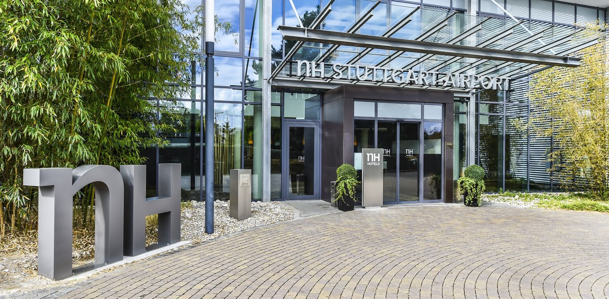 Eingang des NH Stuttgart Airport Hotels mit moderner Glasfassade, großen Buchstaben "nh" und Pflanzen. Der Boden ist mit Pflastersteinen ausgelegt., © NH Hotel Stuttgart Airport