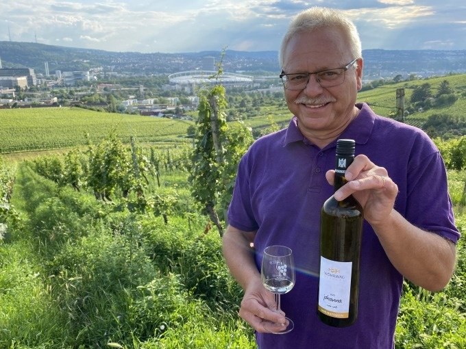 Ein Mann in einem lila Hemd hält eine Weinflasche und ein Glas in einem Weinberg. Im Hintergrund ist Stuttgart mit einem Stadion zu sehen., © Weingenuss Stuttgart Ein Mann in einem lila Hemd hält eine Weinflasche und ein Glas in einem Weinberg. Im Hintergrund ist Stuttgart mit einem Stadion zu sehen., © Weingenuss Stuttgart