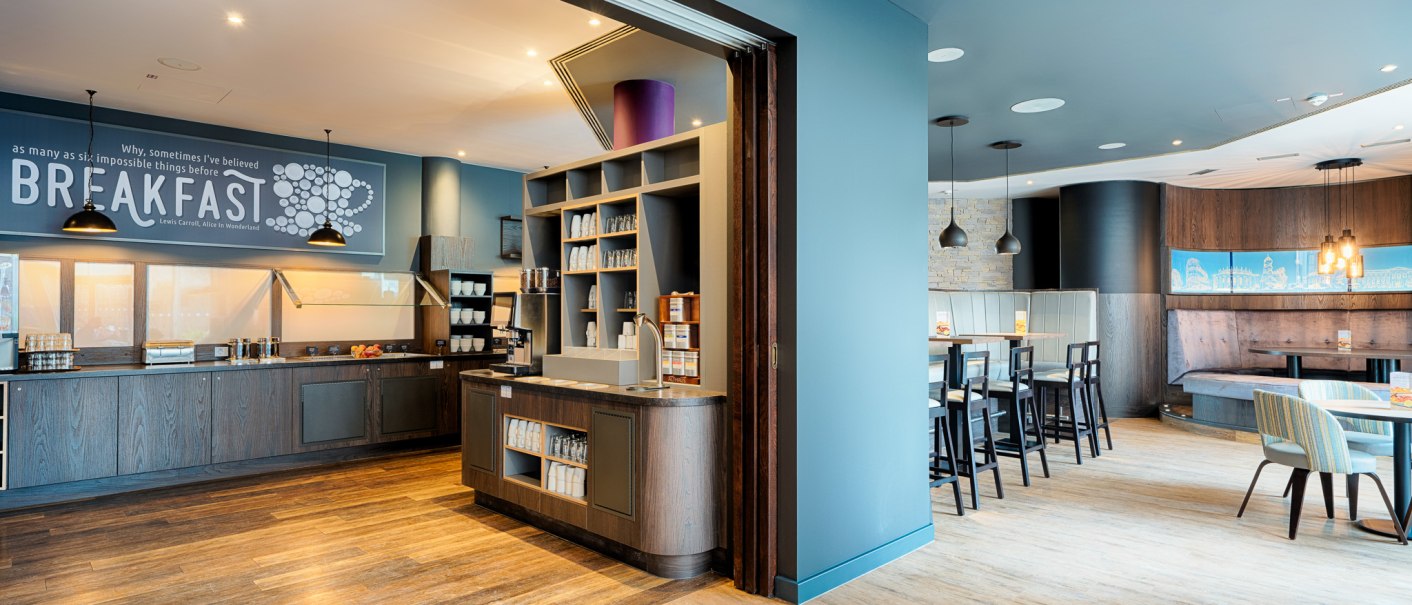 Moderner Fr&uuml;hst&uuml;cksbereich mit Selbstbedienungstheke, Regalen und Sitzgelegenheiten. Helle Holzb&ouml;den und stilvolle Beleuchtung schaffen eine einladende Atmosph&auml;re., &copy; Premier Inn Germany