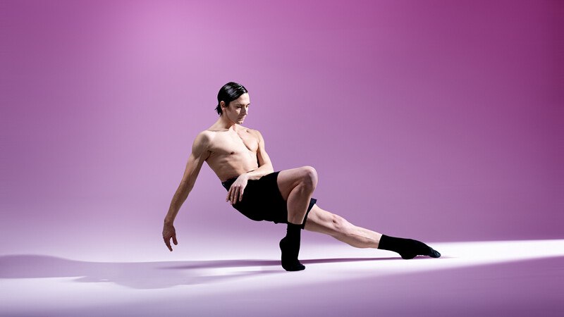 Ein Balletttänzer in schwarzer Kleidung posiert elegant vor einem lila Hintergrund, zeigt Balance und Körperbeherrschung., © Württembergische Staatstheater Stuttgart Ein Balletttänzer in schwarzer Kleidung posiert elegant vor einem lila Hintergrund, zeigt Balance und Körperbeherrschung., © Württembergische Staatstheater Stuttgart