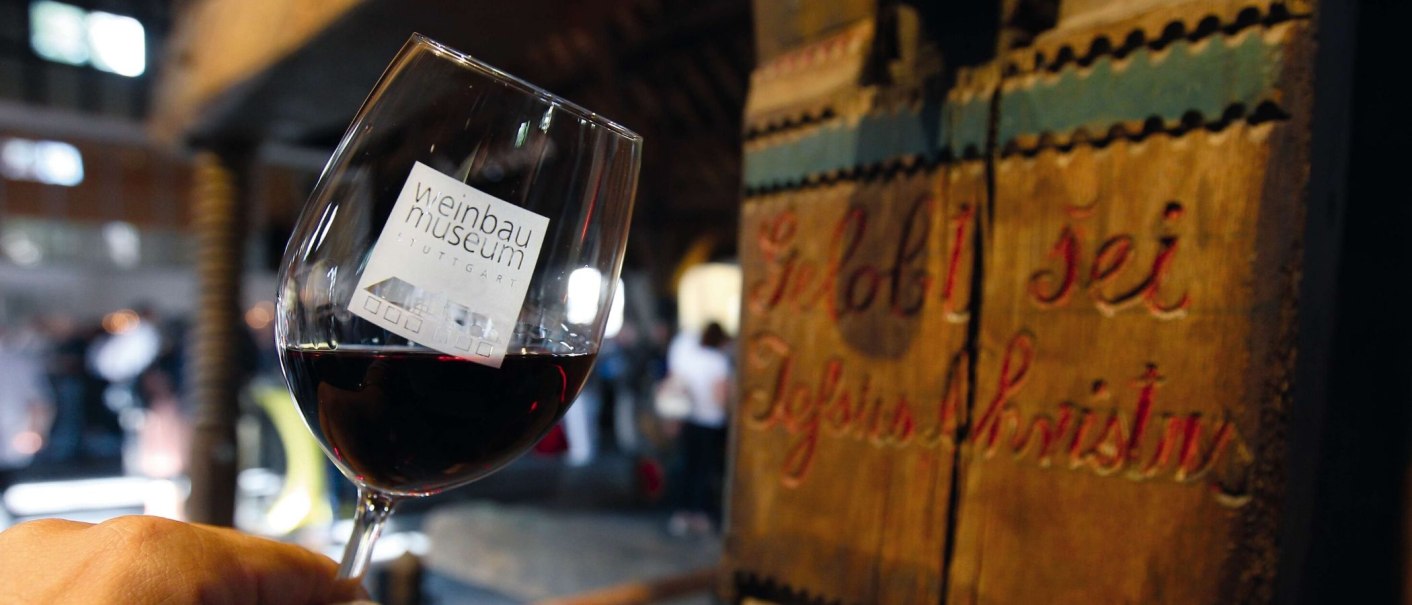 A wine glass with the logo of the Stuttgart Wine Museum is held in front of an old wooden barrel. People can be seen blurred in the background., © Stuttgart-Marketing GmbH, Thomas Niedermüller