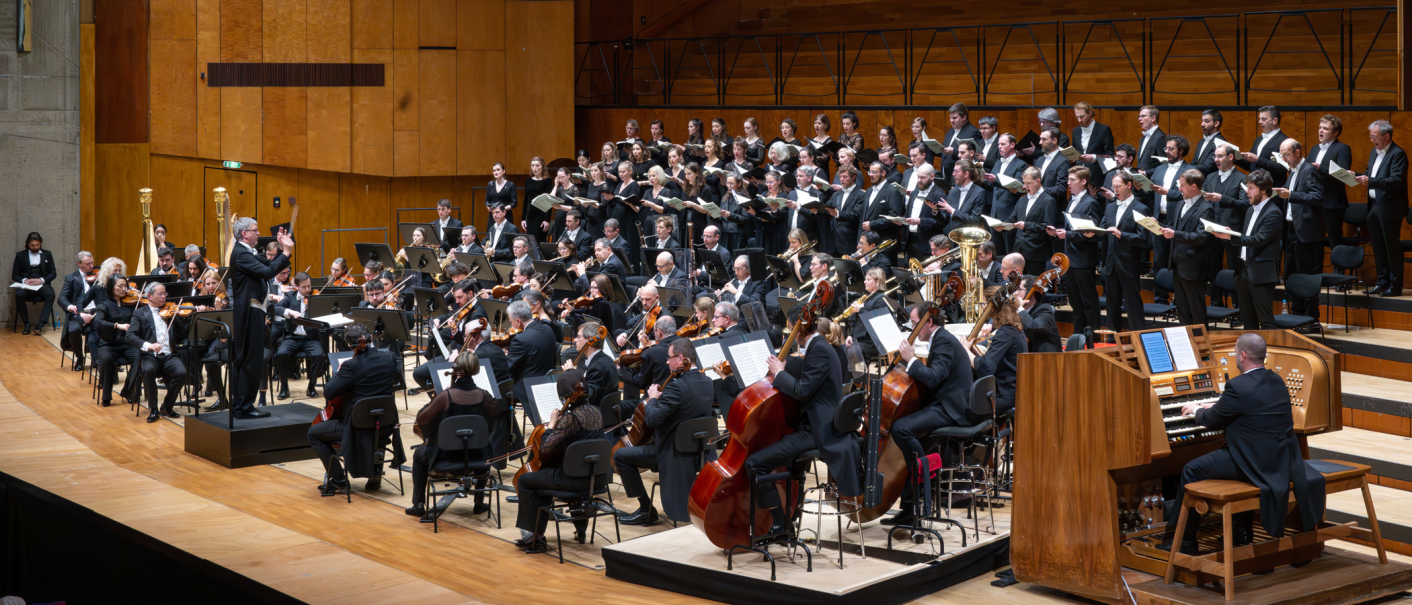 A large orchestra and choir perform in a concert hall. The conductor stands in front of the musicians while an organist plays the organ., &copy; Holger Schneider