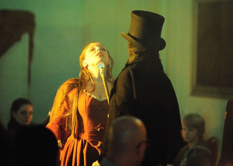 A woman in a red dress stands in front of a man in a black coat and top hat. The scene has a dramatic effect and is held in warm light., © Gruseldinner GmbH & Co. KG A woman in a red dress stands in front of a man in a black coat and top hat. The scene has a dramatic effect and is held in warm light., © Gruseldinner GmbH & Co. KG