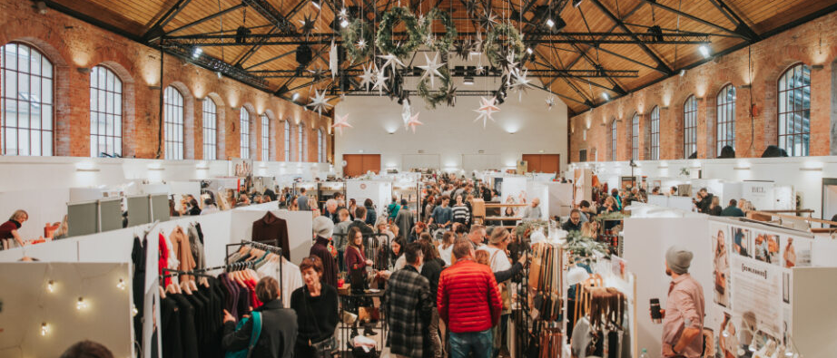 Große Halle mit Weihnachtsmarkt, dekoriert mit Sternen und Kränzen. Viele Menschen schlendern zwischen den Ständen mit Kleidung und Accessoires., © BLICKFANG Messe GmbH