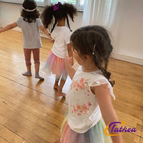 Drei kleine Mädchen tanzen fröhlich in einem Raum mit Holzboden. Sie tragen bunte Röcke und T-Shirts. Im Hintergrund sind Fenster zu sehen., © Tathea - Lena Oschowitzer