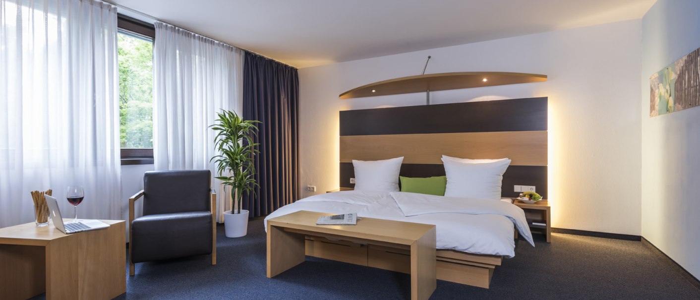 Modernes Hotelzimmer mit Doppelbett, Sessel, Tisch, Laptop und Glas Wein. Fenster mit Vorhängen, Pflanze und stilvolle Beleuchtung., © Hotel Berlin aZIS Hotel BetriebsGmbH