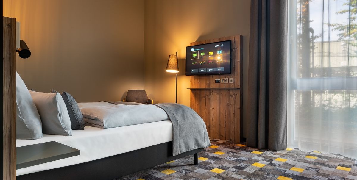 Modernes Hotelzimmer mit einem Bett, einem an der Wand montierten Fernseher und einer Stehlampe. Der Boden hat ein geometrisches Muster., © Novum Hospitality