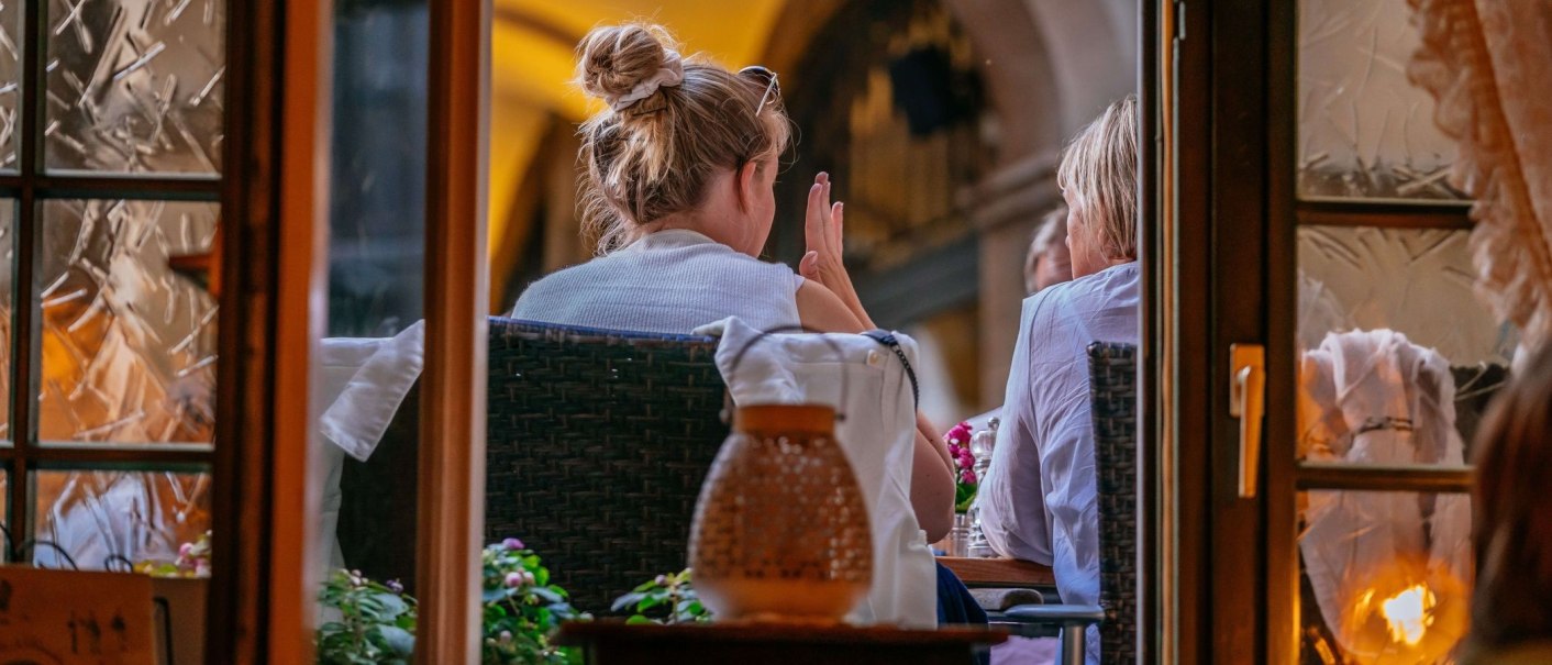Zwei Personen sitzen in einem Caf&eacute;, von au&szlig;en durch ein Fenster gesehen. Eine Person hat einen Dutt, die Atmosph&auml;re wirkt gem&uuml;tlich und einladend., &copy; Thomas Niederm&uuml;ller