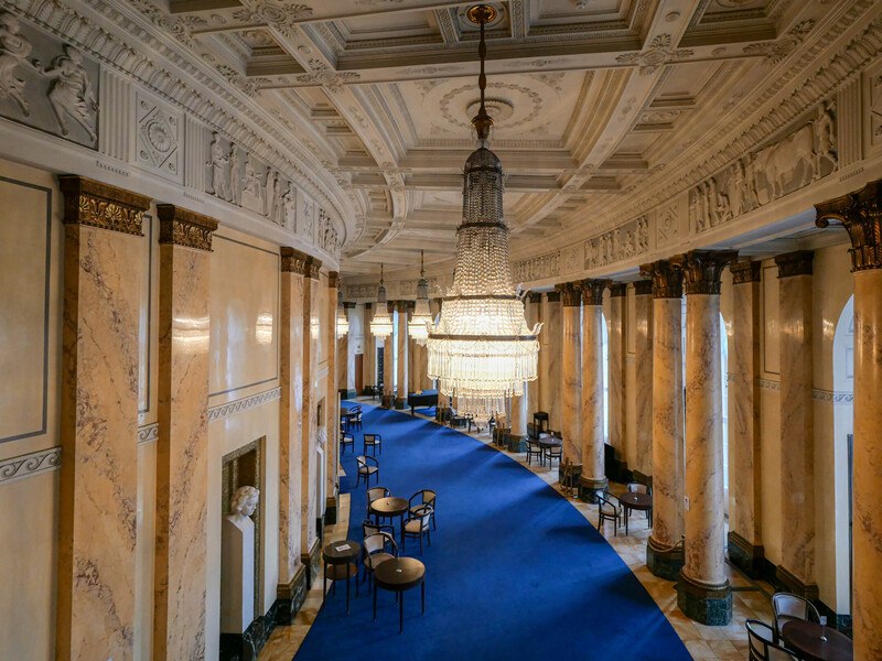 Eleganter Saal mit blauen Teppichen, Marmorsäulen und großen Kronleuchtern, die von der Decke hängen. Tische und Stühle sind entlang des Teppichs angeordnet., © Württembergische Staatstheater Stuttgart Eleganter Saal mit blauen Teppichen, Marmorsäulen und großen Kronleuchtern, die von der Decke hängen. Tische und Stühle sind entlang des Teppichs angeordnet., © Württembergische Staatstheater Stuttgart