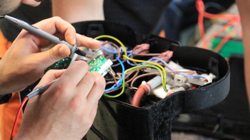 Nahaufnahme einer Person, die mit einem Lötkolben an einer Platine arbeitet, umgeben von bunten Kabeln., © Uprety 2024 Nahaufnahme einer Person, die mit einem Lötkolben an einer Platine arbeitet, umgeben von bunten Kabeln., © Uprety 2024