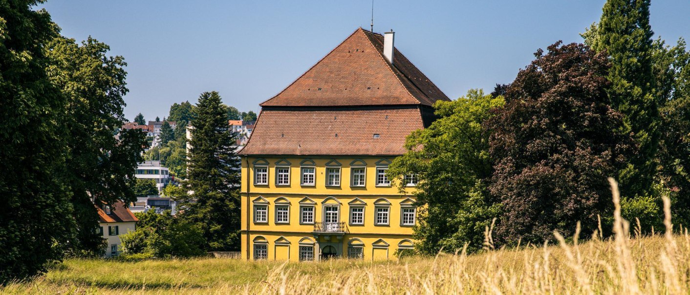 The yellow castle in Wernau stands majestically between green trees and a meadow under a clear sky., © Stuttgart-Marketing GmbH, Sarah Schmid The yellow castle in Wernau stands majestically between green trees and a meadow under a clear sky., © Stuttgart-Marketing GmbH, Sarah Schmid