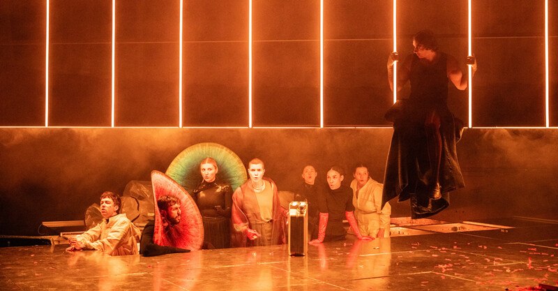 Theaterszene mit Darstellern in fantasievollen Kost&uuml;men auf einer B&uuml;hne. Die Beleuchtung ist orange und erzeugt eine dramatische Atmosph&auml;re., &copy; W&uuml;rttembergische Staatstheater Stuttgart