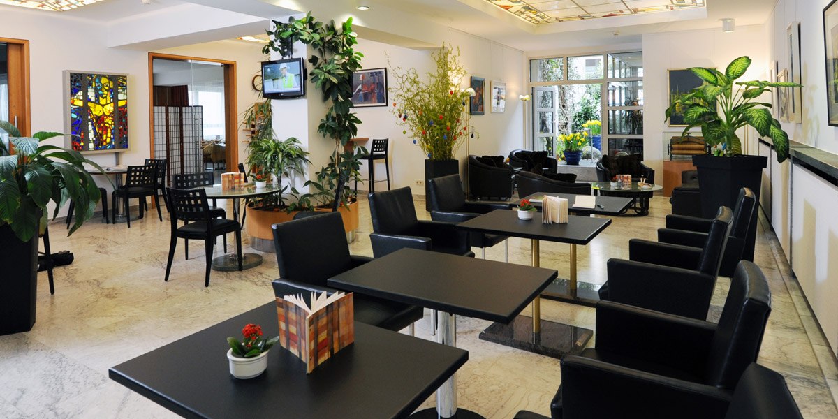 Moderne Lobby mit schwarzen Tischen und Stühlen, Pflanzen und Kunstwerken an den Wänden. Ein Fernseher hängt an der Wand., © Hotel Unger