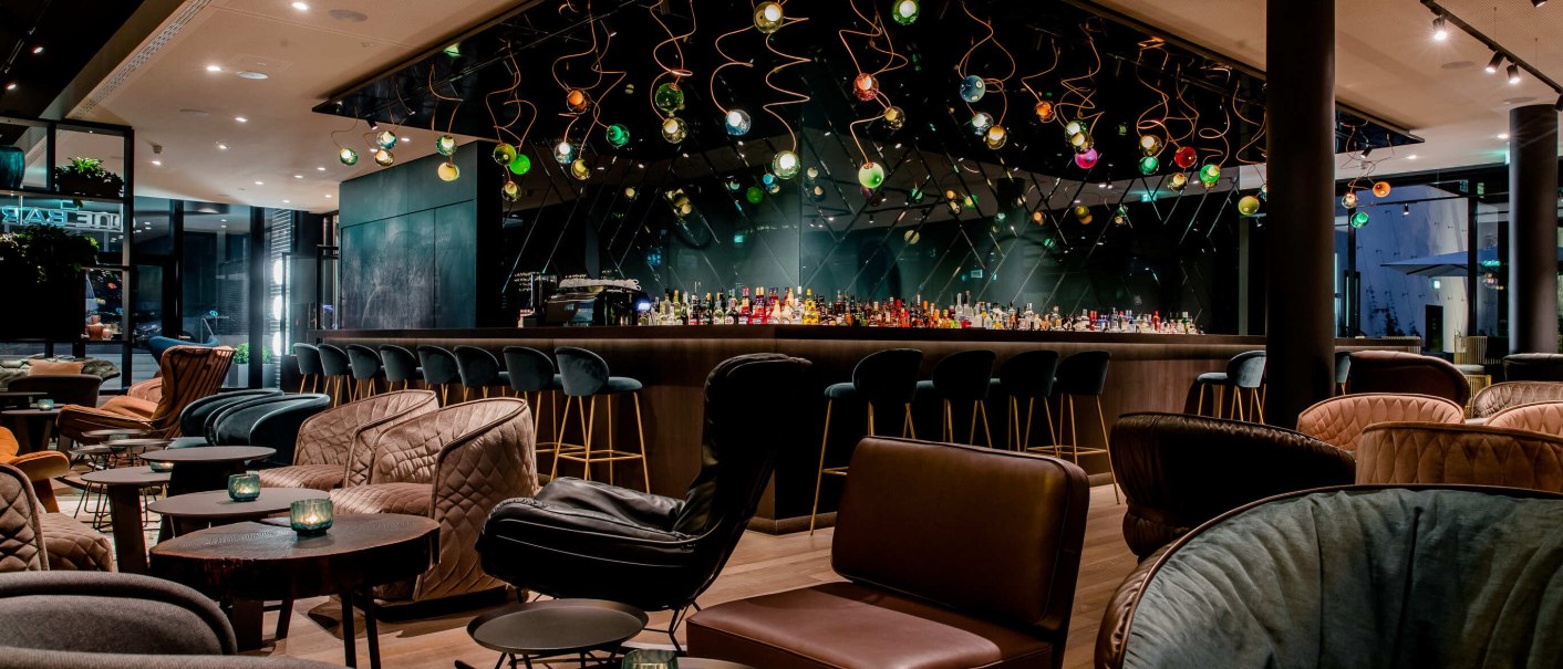 Moderne Bar mit stilvollen Sesseln und Tischen, bunte hängende Lichter und eine lange Theke mit Barhockern., © Motel One - Stuttgart Hauptbahnhof