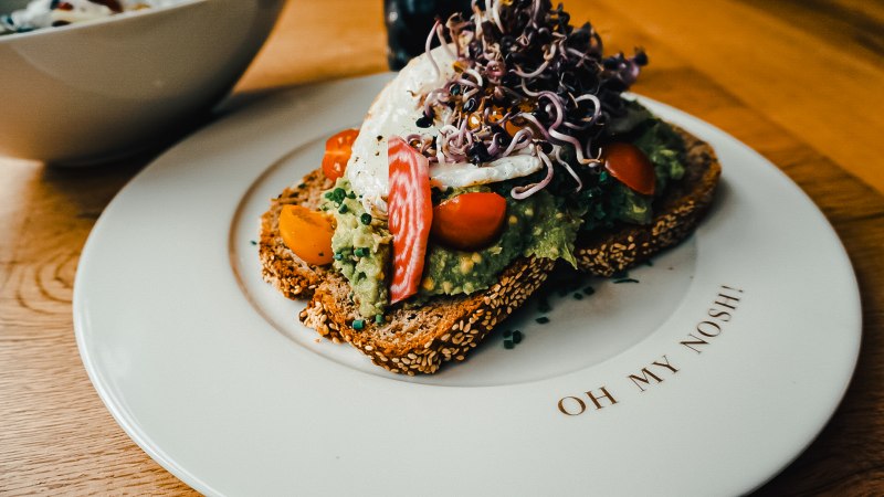 Belegtes Brot mit Avocado, Tomaten und Sprossen auf einem Teller mit der Aufschrift 'Oh my Nosh'., © The Gardeners Nosh