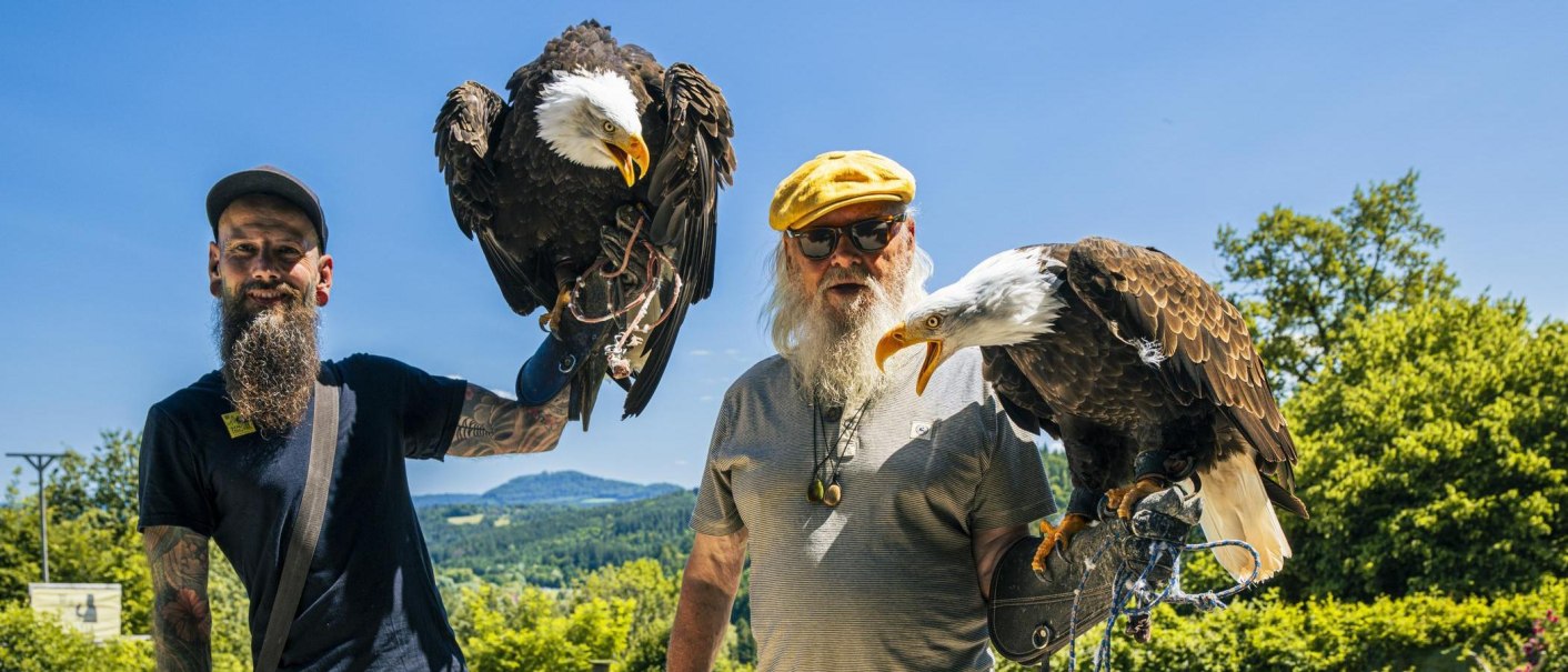 Two men proudly hold bald eagles in their arms. In the background, a green landscape can be seen beneath a clear blue sky., © SMG Stuttgart Marketing GmbH - Sarah Schmid Two men proudly hold bald eagles in their arms. In the background, a green landscape can be seen beneath a clear blue sky., © SMG Stuttgart Marketing GmbH - Sarah Schmid