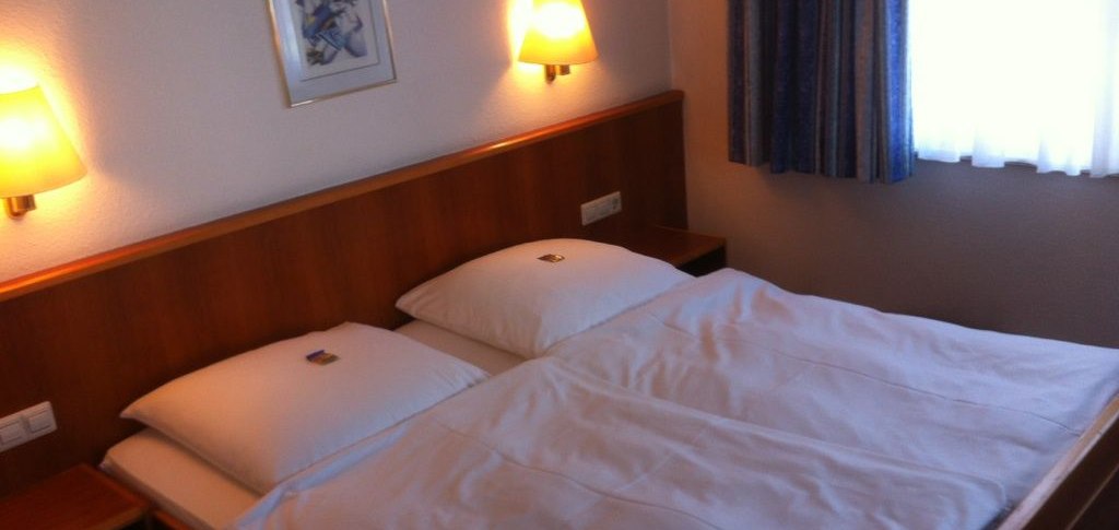 Ein gem&uuml;tliches Doppelzimmer mit einem gro&szlig;en Bett, zwei Wandlampen und einem Bild an der Wand. Ein Fenster mit Vorh&auml;ngen ist zu sehen., &copy; Hotel Sonne garni