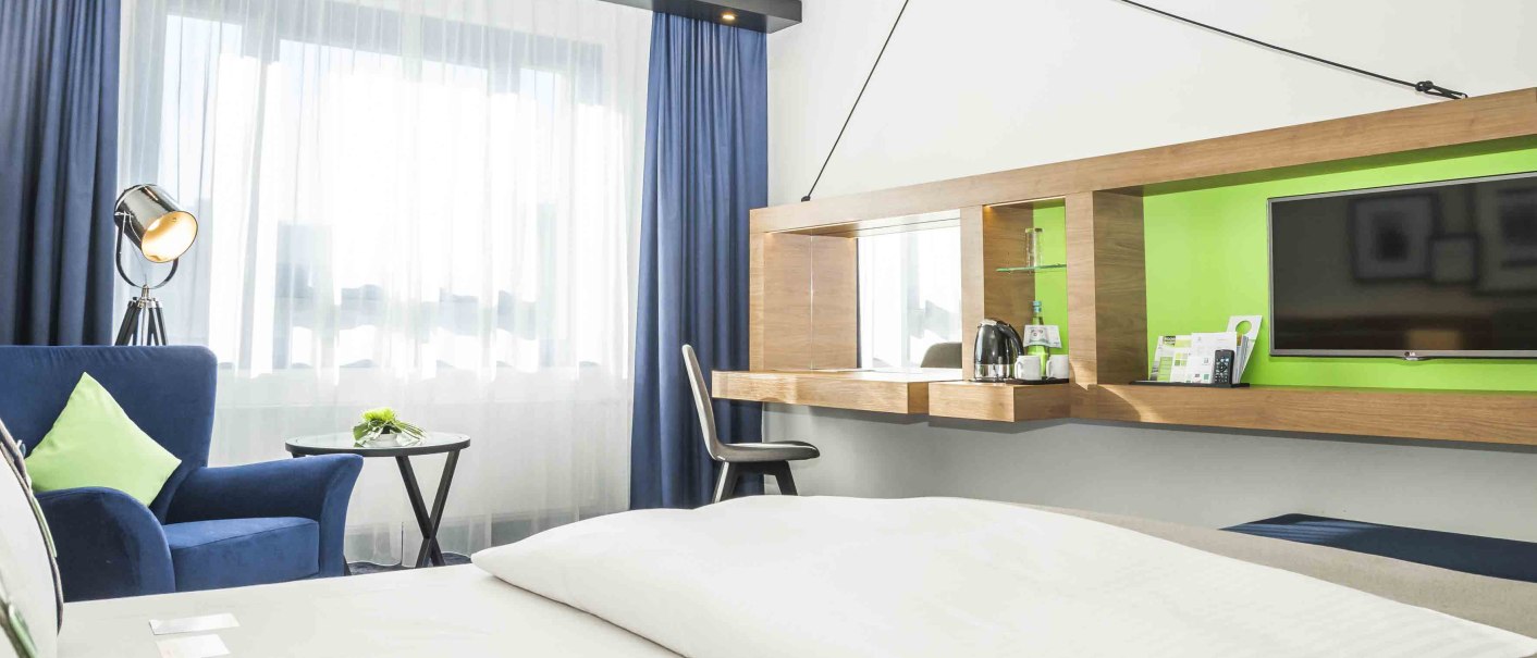 Modern hotel room with white bed, blue armchair, desk, TV and large window front with blue curtains., © Chandler Photography Modern hotel room with white bed, blue armchair, desk, TV and large window front with blue curtains., © Chandler Photography
