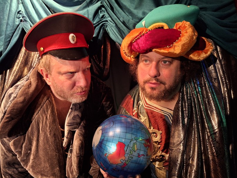 Zwei Männer in fantasievollen Kostümen, einer mit roter Mütze, der andere mit buntem Turban, halten einen Globus. Der Hintergrund ist mit Stoff drapiert., © Theater am Olgaeck Zwei Männer in fantasievollen Kostümen, einer mit roter Mütze, der andere mit buntem Turban, halten einen Globus. Der Hintergrund ist mit Stoff drapiert., © Theater am Olgaeck