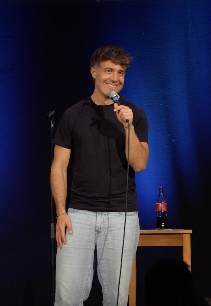 Ein Mann auf einer Bühne hält ein Mikrofon und lächelt. Hinter ihm ein blauer Vorhang und ein Tisch mit einer Flasche., © Theaterhaus Stuttgart e.V. Ein Mann auf einer Bühne hält ein Mikrofon und lächelt. Hinter ihm ein blauer Vorhang und ein Tisch mit einer Flasche., © Theaterhaus Stuttgart e.V.