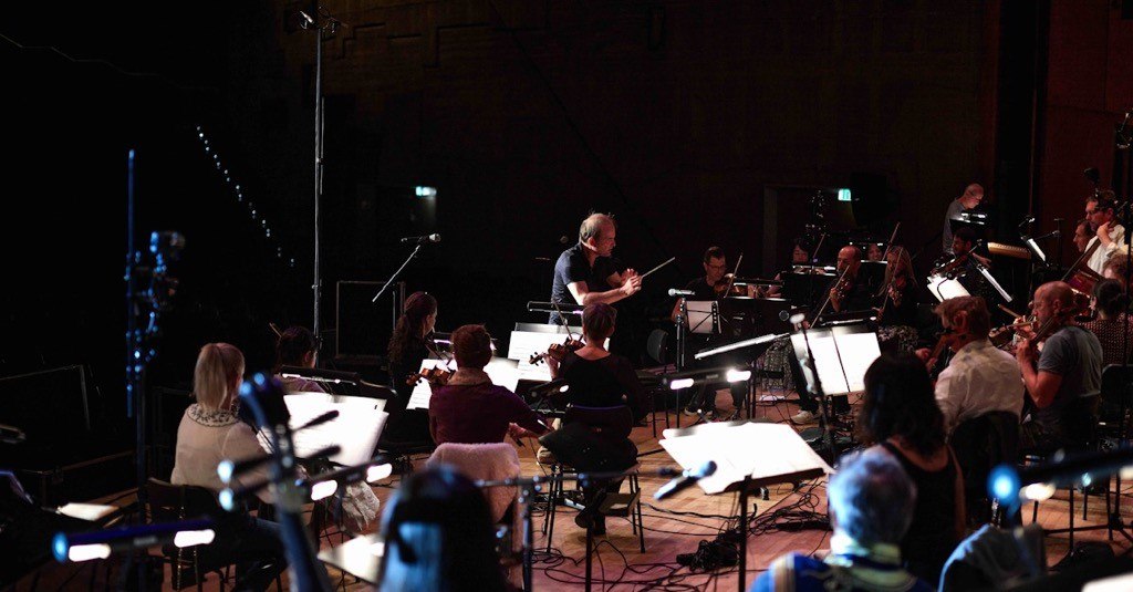 Ein Orchester probt auf einer B&uuml;hne, dirigiert von einem Dirigenten. Musiker spielen verschiedene Instrumente, umgeben von Notenst&auml;ndern und Mikrofonen., &copy; Marc Sch&auml;fer