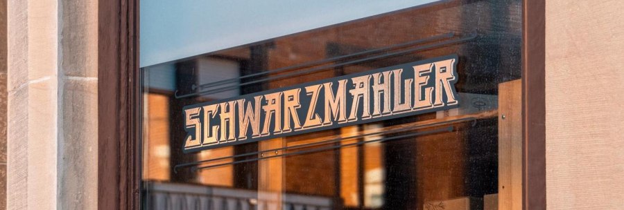 Ein Schaufenster mit dem Schriftzug 'Schwarzmahler' in stilisierter Schrift. Die Umgebung spiegelt sich im Glas., © SMG, Sarah Schmid Ein Schaufenster mit dem Schriftzug 'Schwarzmahler' in stilisierter Schrift. Die Umgebung spiegelt sich im Glas., © SMG, Sarah Schmid