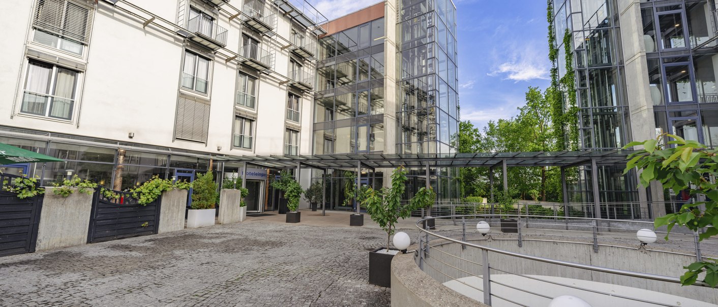 Modern building with glass façade, plants in the entrance area and paved forecourt. Clear lines and lots of glass characterize the design., © PLAZA Hotelgroup GmbH