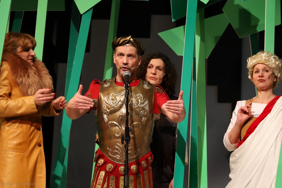 Vier Schauspieler in historischen Kostümen auf einer Bühne. Einer trägt eine römische Rüstung, die anderen sind in antiker und moderner Kleidung gekleidet., © Sabine Haymann Vier Schauspieler in historischen Kostümen auf einer Bühne. Einer trägt eine römische Rüstung, die anderen sind in antiker und moderner Kleidung gekleidet., © Sabine Haymann