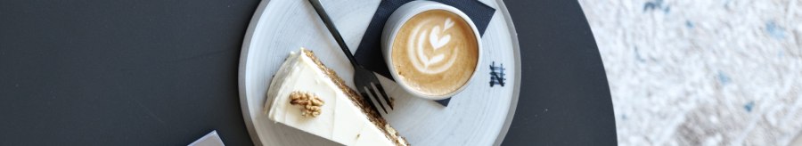 Latte mit Herz-Latte-Art und ein Stück Kuchen auf einem Teller auf schwarzem Tisch, daneben eine Karte mit „home interior“., © Taraba Latte mit Herz-Latte-Art und ein Stück Kuchen auf einem Teller auf schwarzem Tisch, daneben eine Karte mit „home interior“., © Taraba