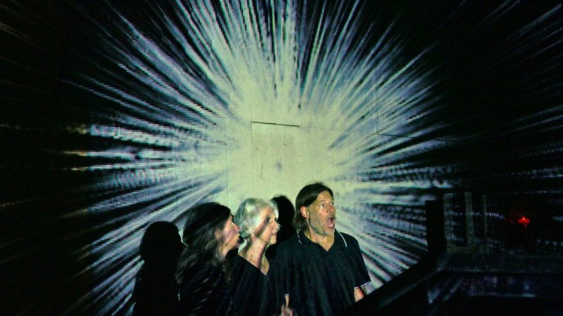 Drei Personen stehen vor einer strahlenf&ouml;rmigen Projektion, die von einem zentralen Punkt ausgeht. Die Szene wirkt dramatisch und k&uuml;nstlerisch., &copy; Diethard Verleger