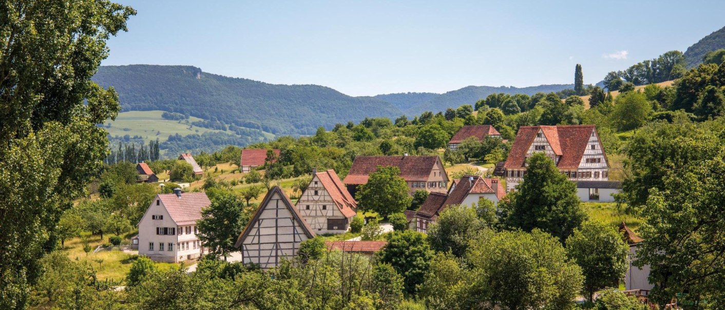 A picturesque village with traditional half-timbered houses, surrounded by green hills and trees, under a clear blue sky., © © Stuttgart-Marketing GmbH A picturesque village with traditional half-timbered houses, surrounded by green hills and trees, under a clear blue sky., © © Stuttgart-Marketing GmbH