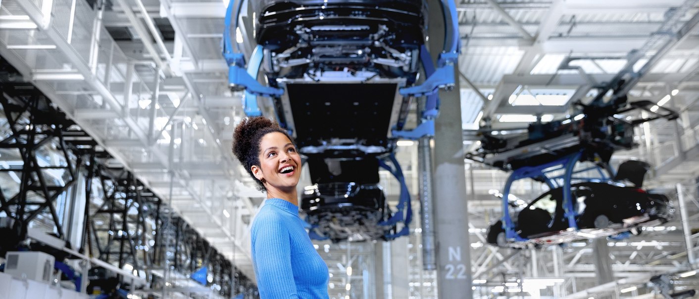 Woman in blue sweater in modern car factory with hanging car bodies in the background., © Mercedes-Benz AG